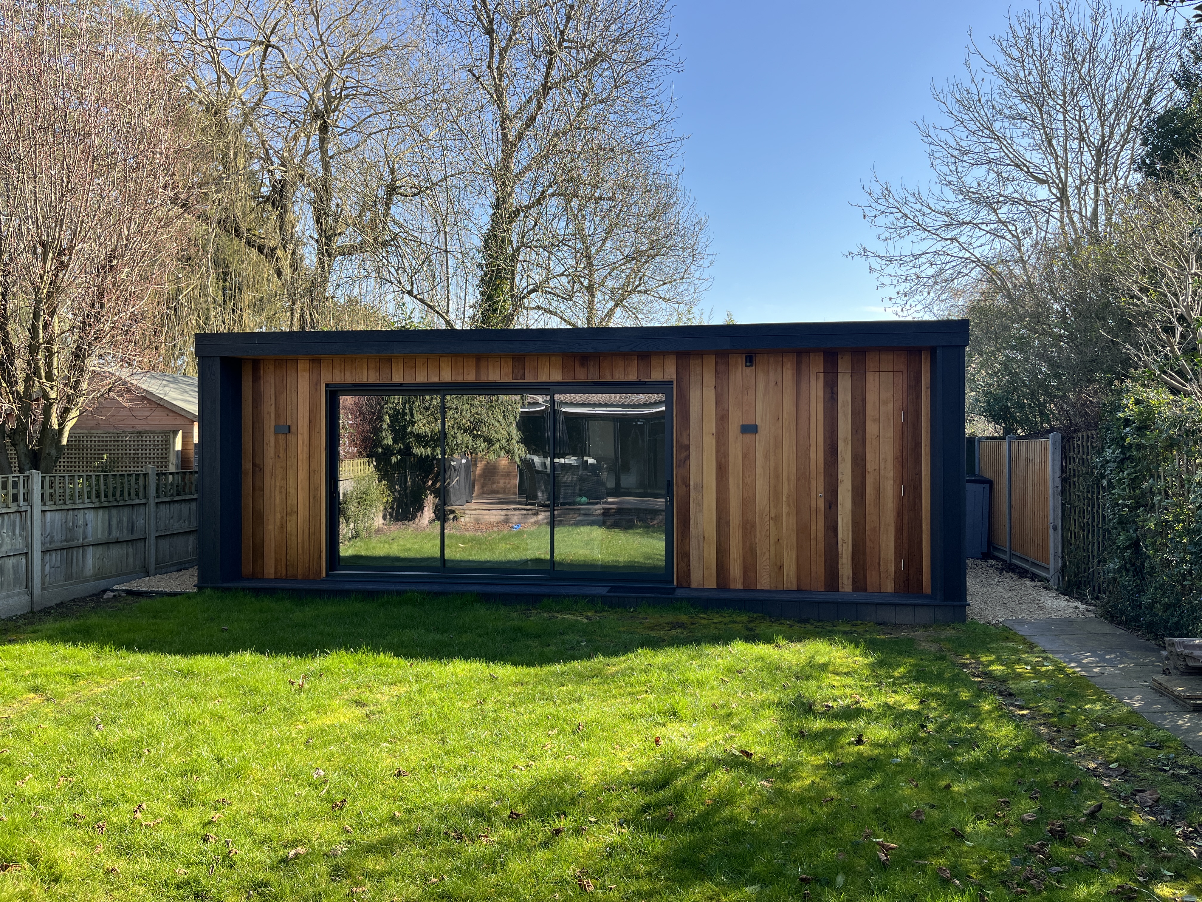 Rustic wooden garden room with large glass doors, featuring a covered hot tub area and spacious deck, surrounded by lush greenery and trees.