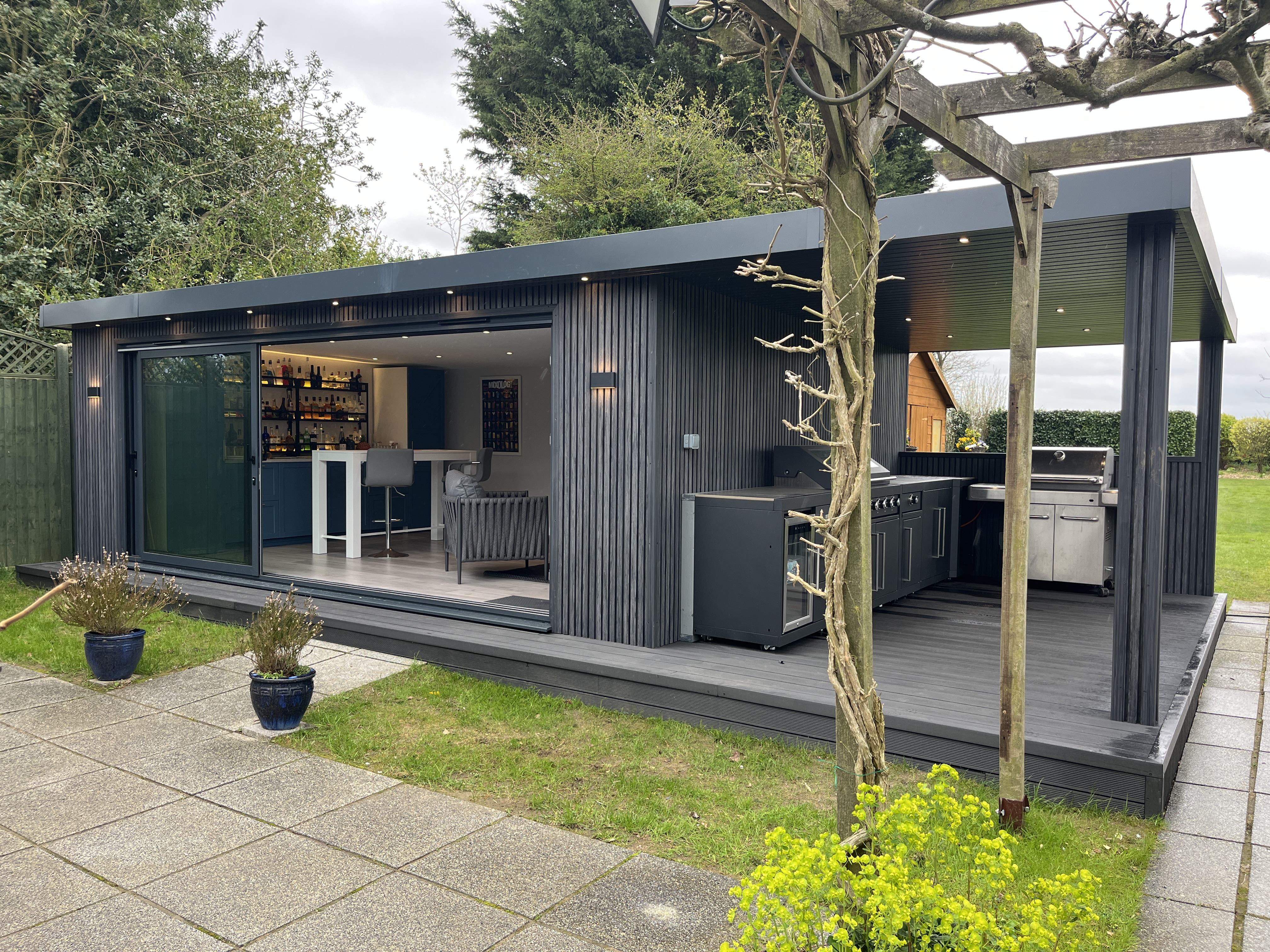 Modern black garden studio with sleek glass doors and minimalist design, featuring a spacious deck and lush green lawn, surrounded by vibrant foliage.