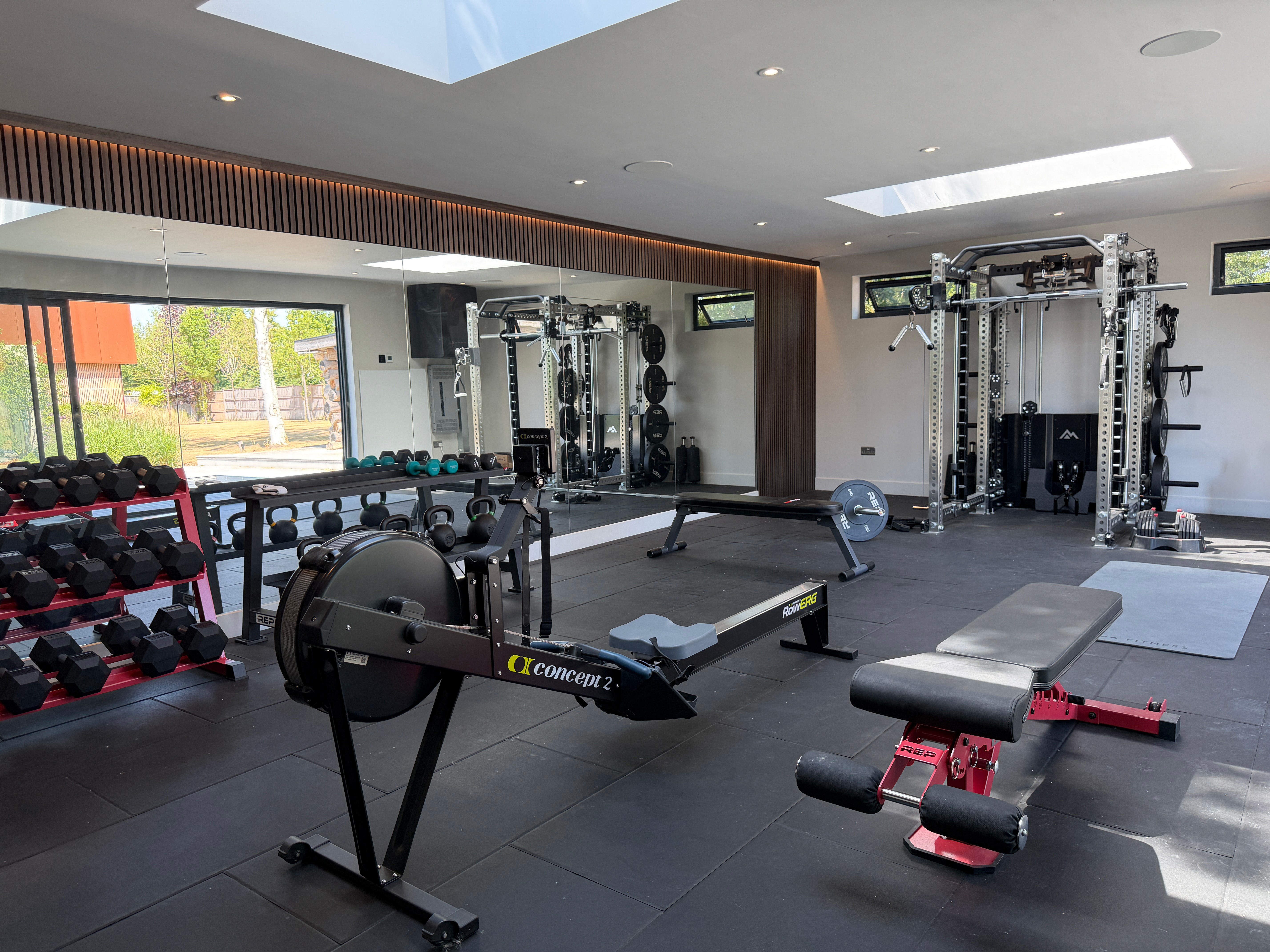 Modern home gym with open glass doors, featuring exercise equipment, weights, and a sleek wooden interior, set within a lush garden environment.