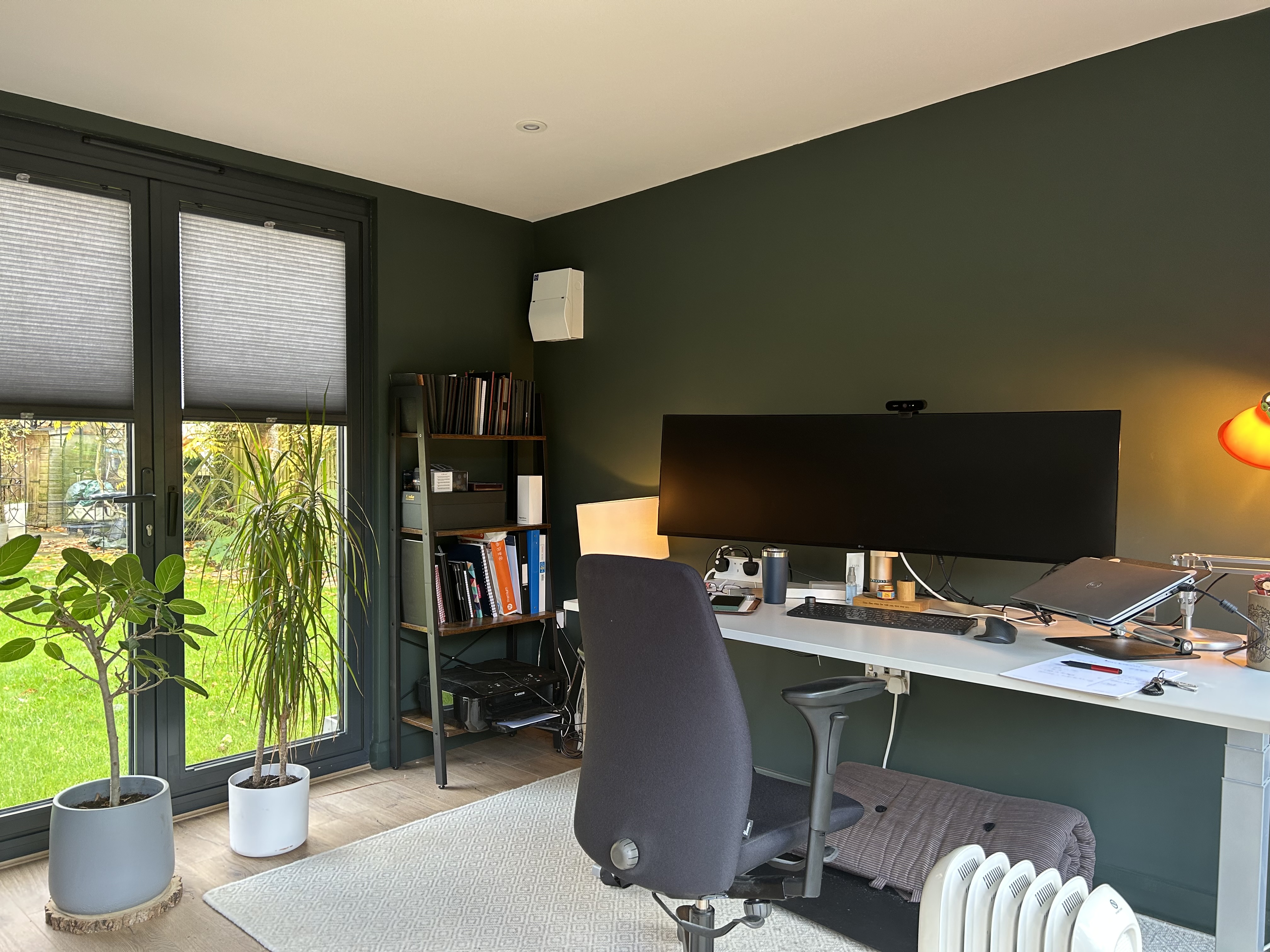 Contemporary garden office with bi-fold glass doors, featuring a comfortable workspace with a desk, ergonomic chair, exercise bike, and stylish decor, set on a paved patio with adjacent greenery.