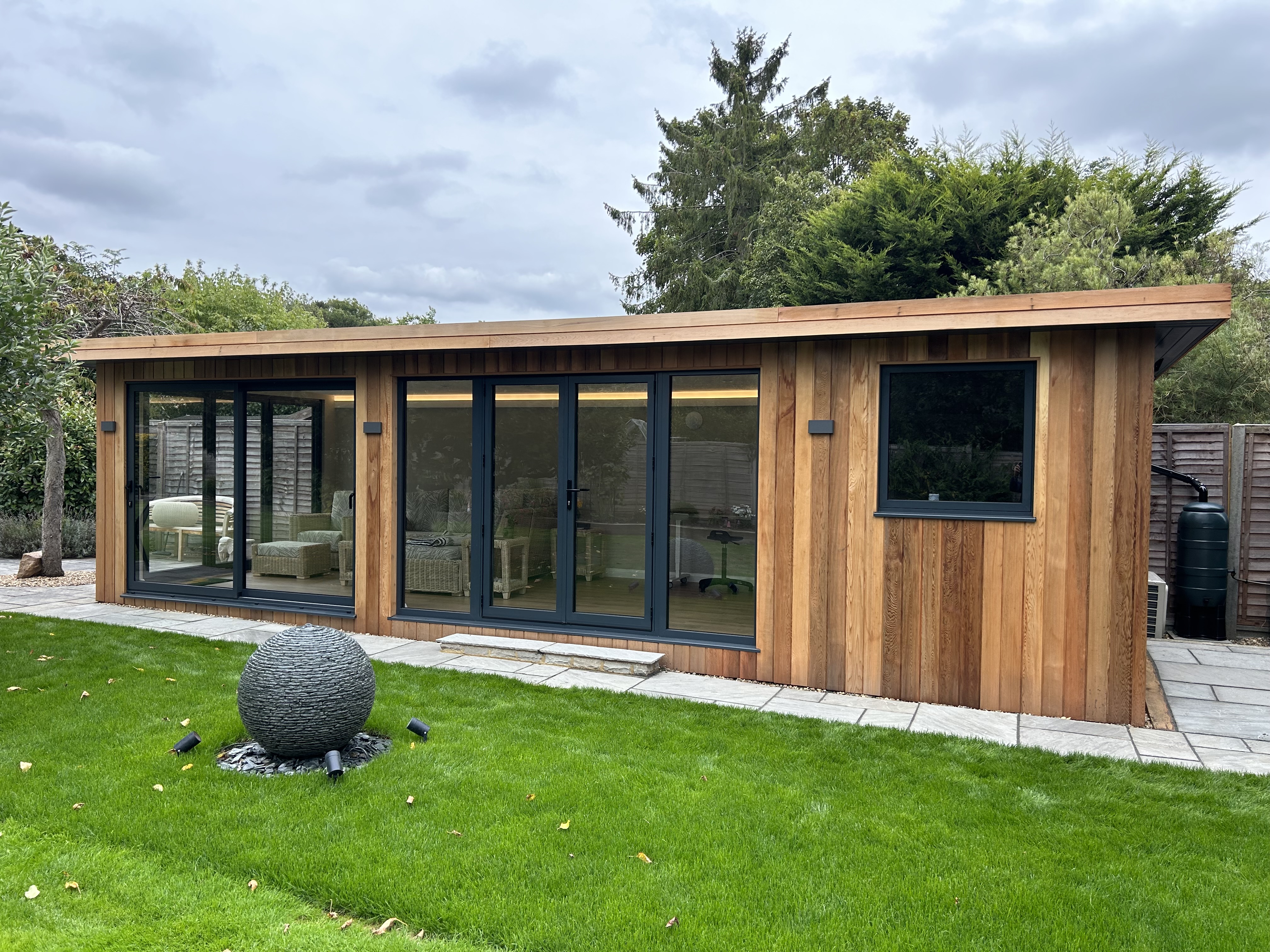 Modern garden room with large glass doors and wood cladding, featuring a comfortable interior and surrounded by a well-manicured lawn with a decorative stone sphere.