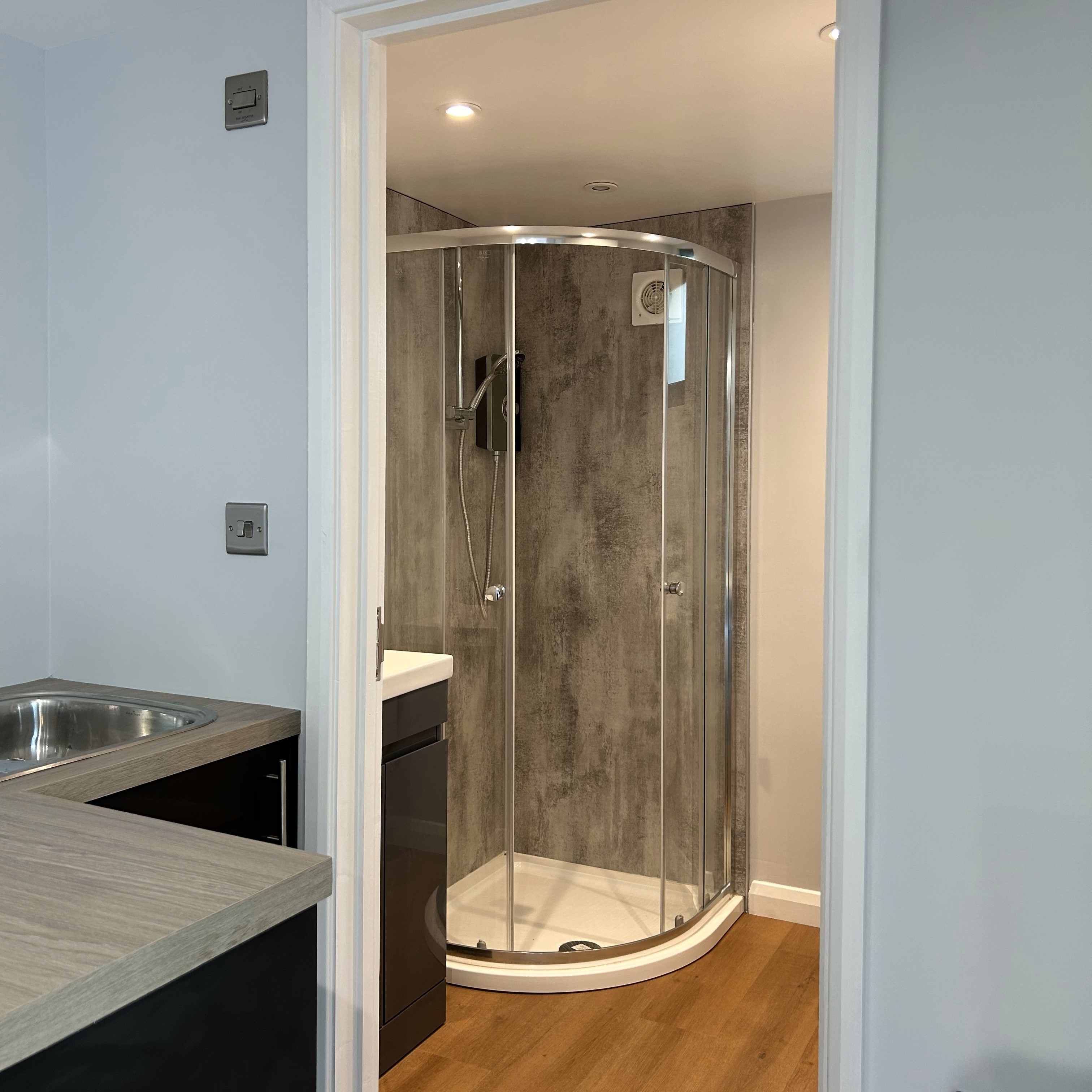 The compact bathroom features a modern shower enclosure with grey textured walls, a sleek glass door, and a white toilet positioned under a small, frosted window that provides natural light while maintaining privacy. An efficient exhaust fan is installed above the shower area.