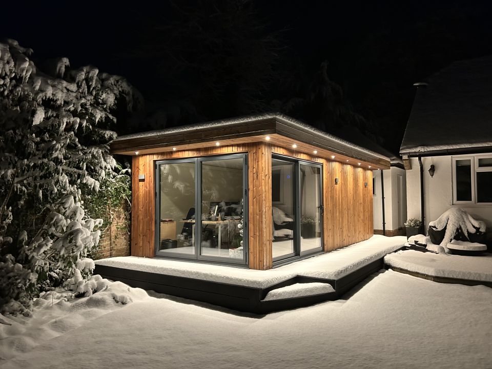 Modern wooden garden room illuminated at night, featuring large glass doors and a snow-covered deck, creating a warm and inviting atmosphere amidst a serene winter landscape.