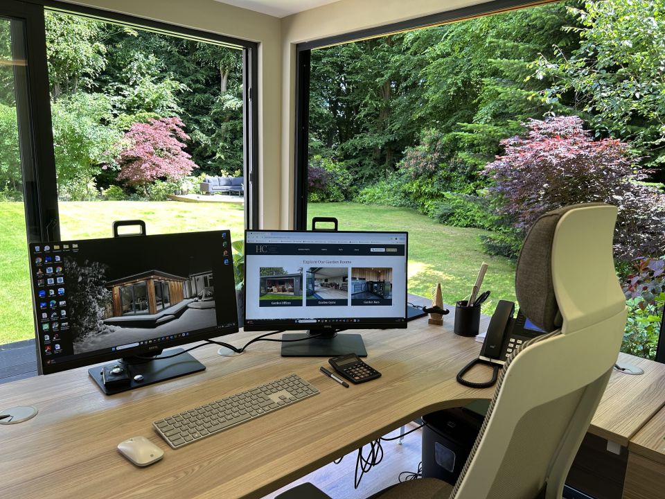 Modern home office with large windows, featuring a dual-monitor setup on a wooden desk, ergonomic chair, and a beautiful view of a lush, green garden with colourful foliage.