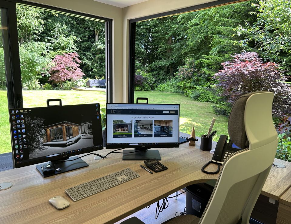 Modern home office with large windows, featuring a dual-monitor setup on a wooden desk, ergonomic chair, and a beautiful view of a lush, green garden with colourful foliage.