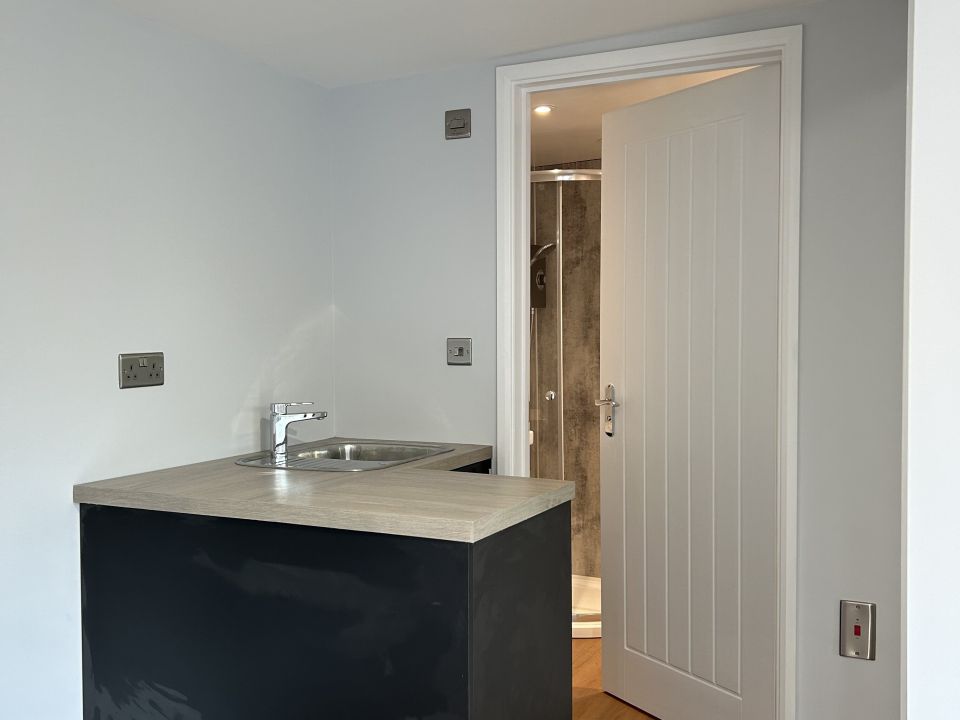 Modern and minimalist kitchenette featuring a sleek black cabinet with a wooden countertop and a stainless steel sink, adjacent to a doorway leading to a bathroom with a contemporary shower.