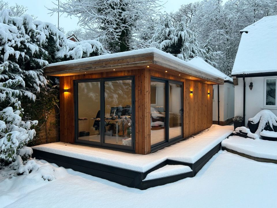 Cosy wooden garden room with large glass doors, warmly lit exterior lights, and surrounded by a picturesque snow-covered landscape, creating a serene winter retreat.