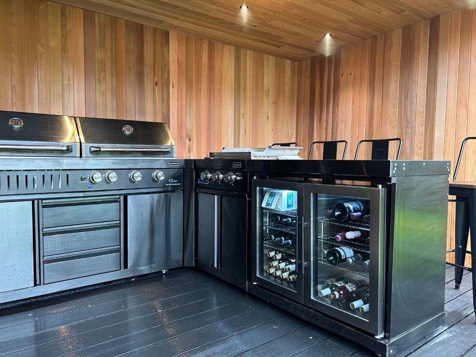 The modern outdoor kitchen showcases a sleek, stainless steel BBQ setup featuring multiple grills and control knobs. Adjacent is a stylish black bar area, complete with a built-in wine cooler stocked with various bottles. The warm, wooden paneling of the walls and ceiling, combined with spot lighting, creates an inviting atmosphere for cooking and entertaining. High bar stools add a comfortable seating option for guests.
