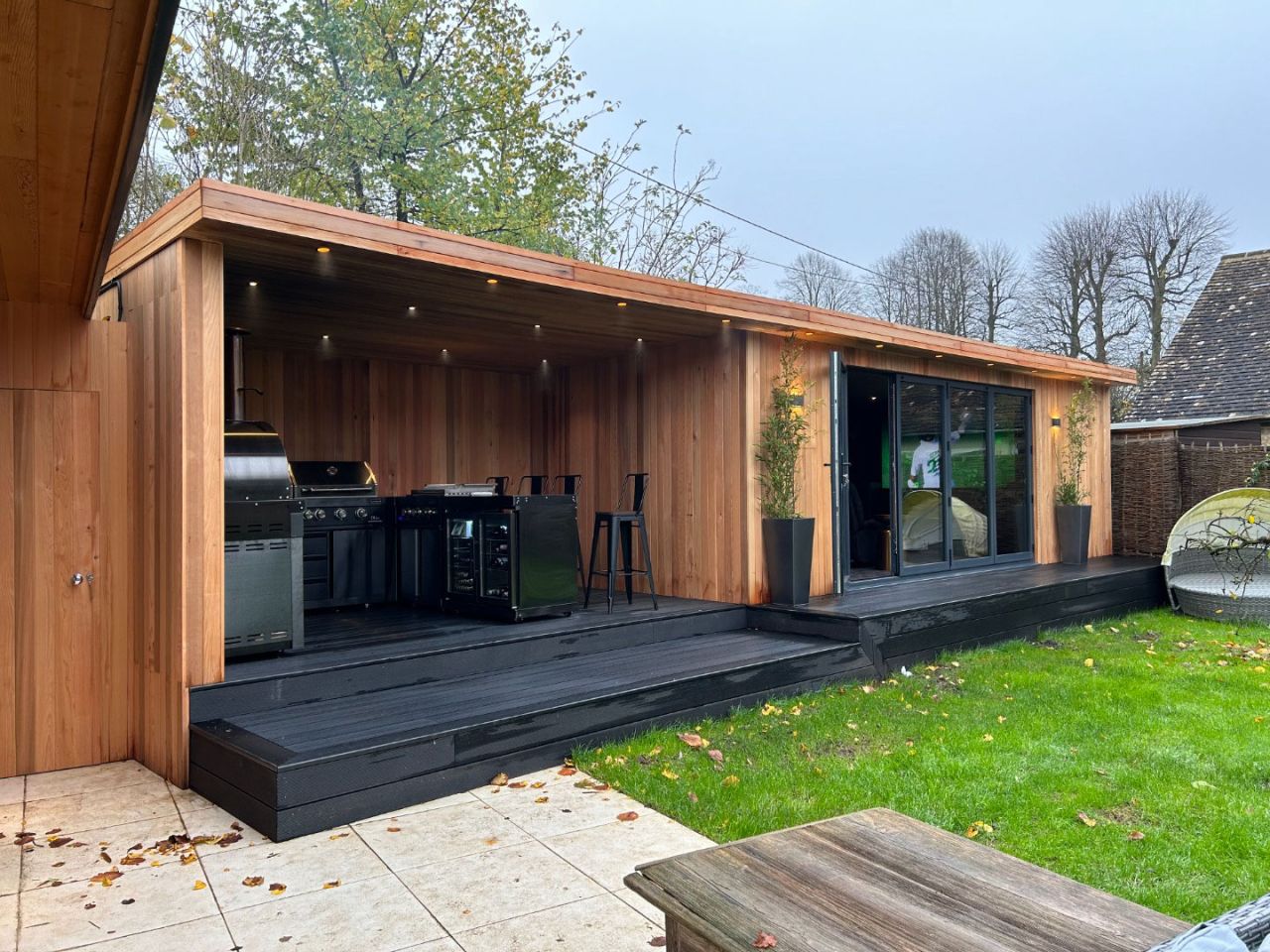 Stylish outdoor kitchen and entertainment area with wood cladding, featuring a barbecue grill, bar seating, and sliding glass doors, surrounded by a lush garden setting.