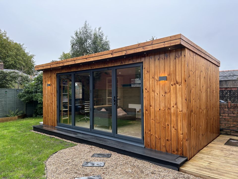 Cosy wooden garden office with large glass sliding doors, warm interior lighting, and a charming gravel pathway, nestled in a lush garden setting.