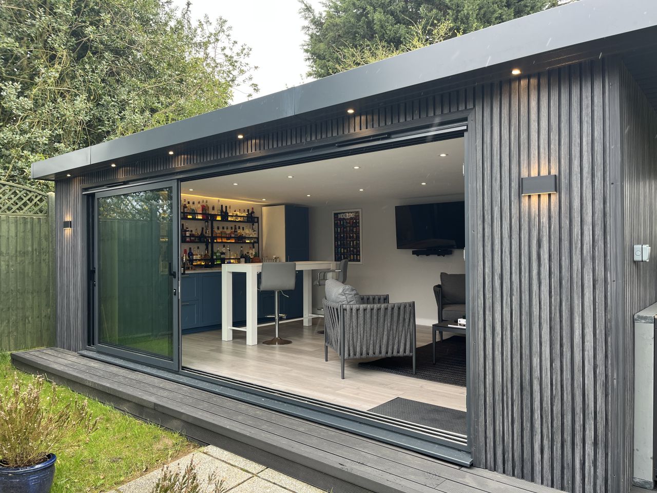 Stylish outdoor kitchen and entertainment area with wood cladding, featuring a barbecue grill, bar seating, and sliding glass doors, surrounded by a lush garden setting.
