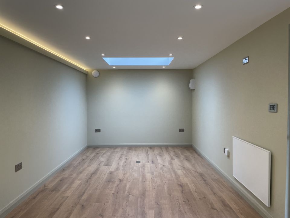 Modern and minimalist interior of a garden room, featuring light wood flooring, recessed lighting, a skylight, and soft beige walls, creating a bright and airy space.