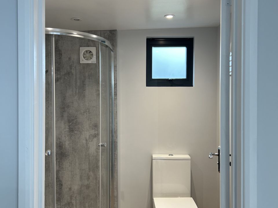 The compact bathroom features a modern shower enclosure with grey textured walls, a sleek glass door, and a white toilet positioned under a small, frosted window that provides natural light while maintaining privacy. An efficient exhaust fan is installed above the shower area.