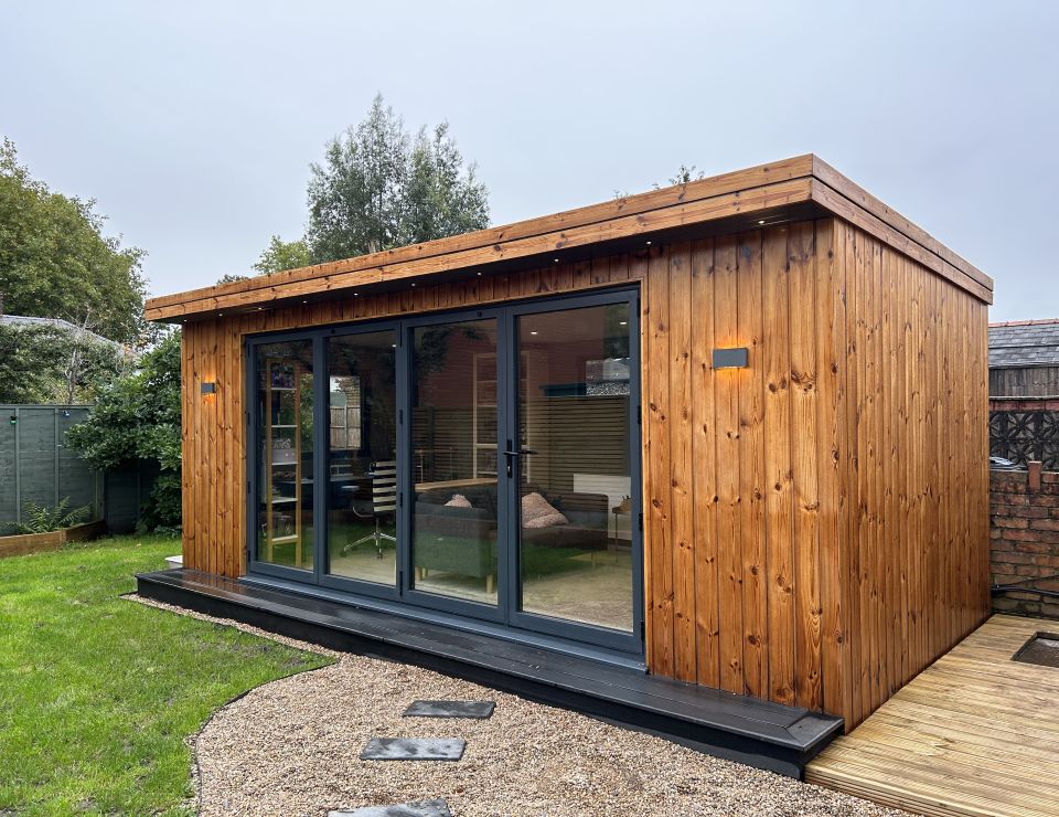 Cosy wooden garden office with large glass sliding doors, warm interior lighting, and a charming gravel pathway, nestled in a lush garden setting.