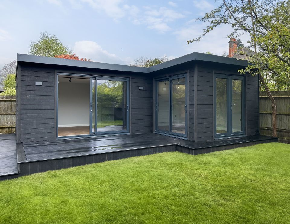 Modern garden room with bi-fold glass doors, featuring a cosy, rustic interior with comfortable seating, a wood-burning stove, and an adjacent covered patio, set within a lush green garden.