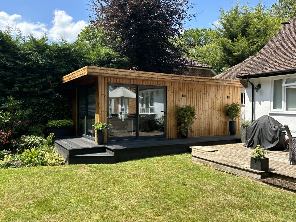 Charming wooden garden room with large glass doors, surrounded by lush greenery, featuring a cosy deck and adjacent patio area, creating an inviting outdoor living space.