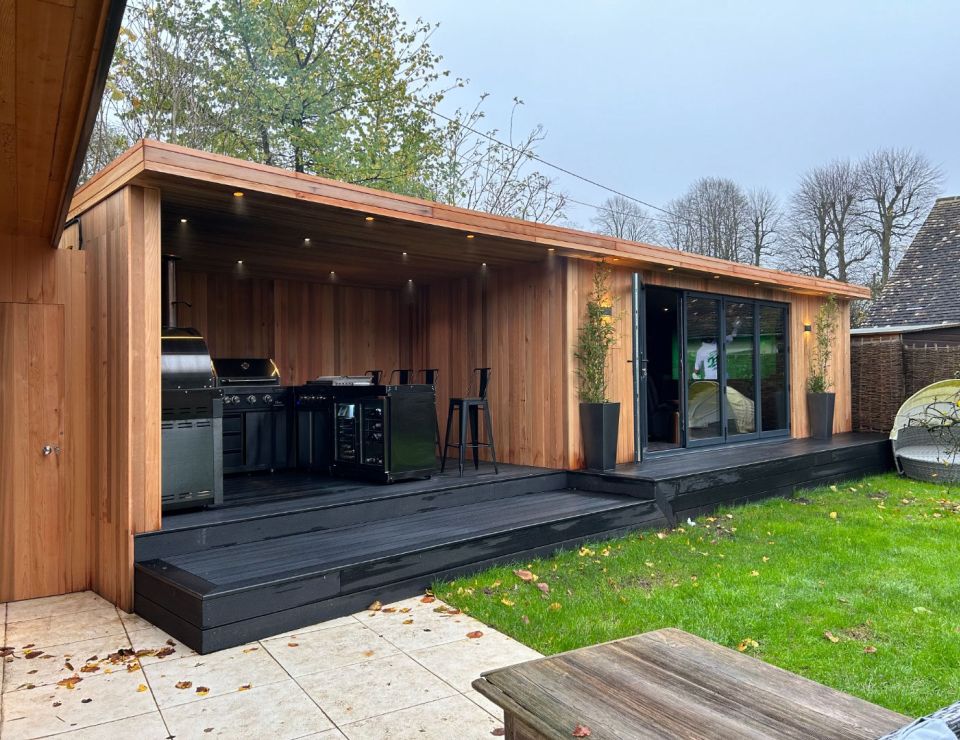 Elegant outdoor kitchen and entertainment space with wooden cladding, featuring a barbecue grill, bar seating, and sliding glass doors, set within a vibrant garden environment.