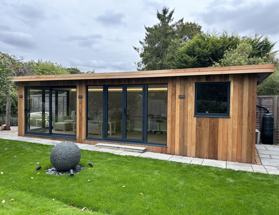 Cosy garden room with wooden cladding and black-framed windows and doors, featuring a small wooden deck with built-in planters, set in a well-kept garden.