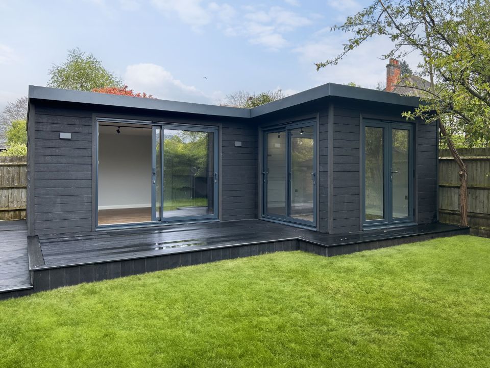 Sleek black garden room with large sliding glass doors and a wrap-around deck, set within a lush green lawn and fenced garden, offering a modern and tranquil outdoor living space.